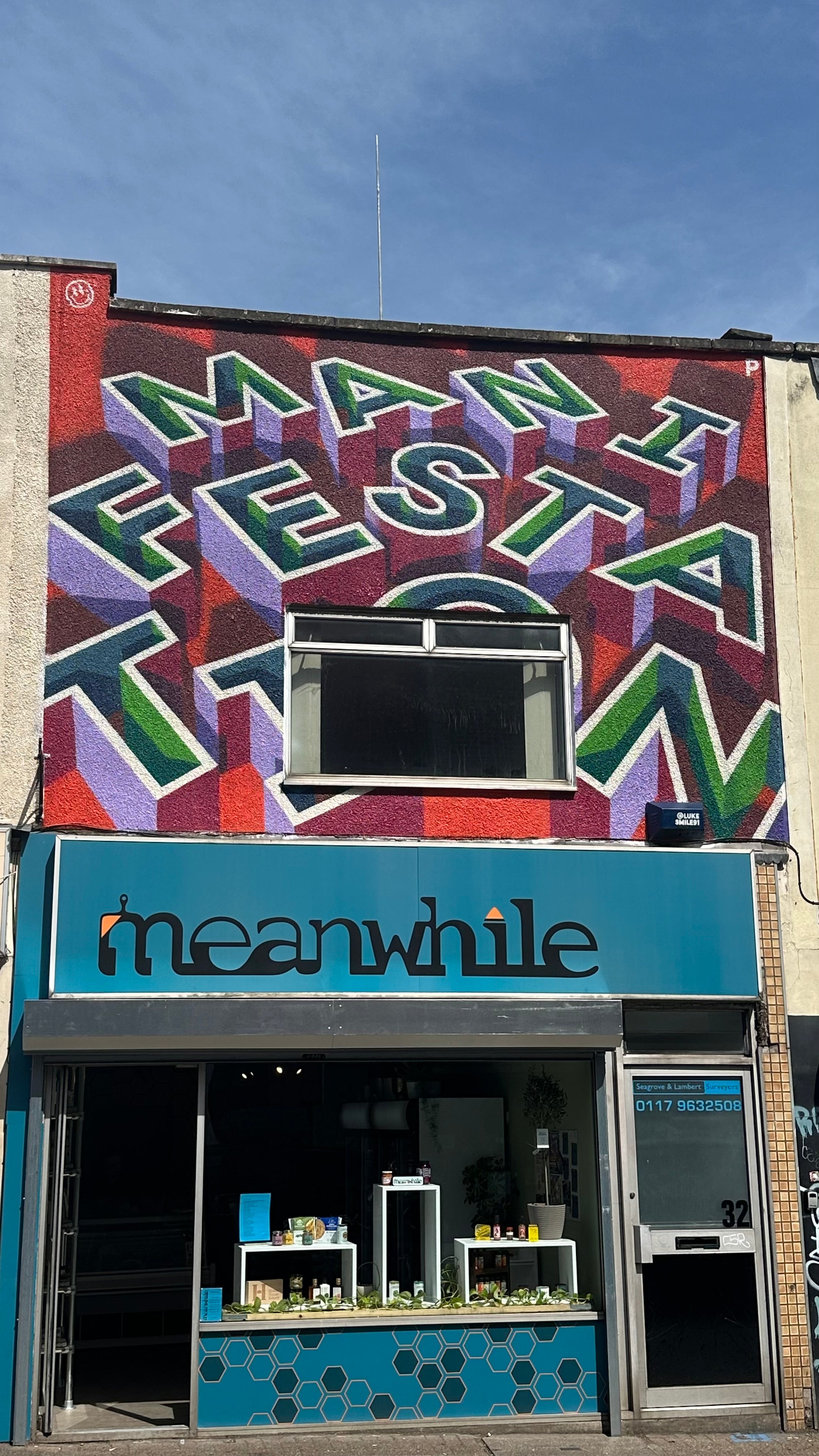 A storefront facade with a large colorful mural reading 'MANIFEST' across the upper wall and a turquoise ' meanwhile' sign above a glass-front entrance and counter.